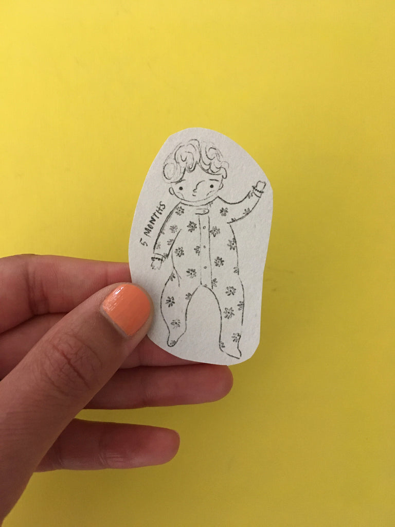 Meenal's hand holding a small cut-out sketch of a baby against a bright sunshine yellow background.