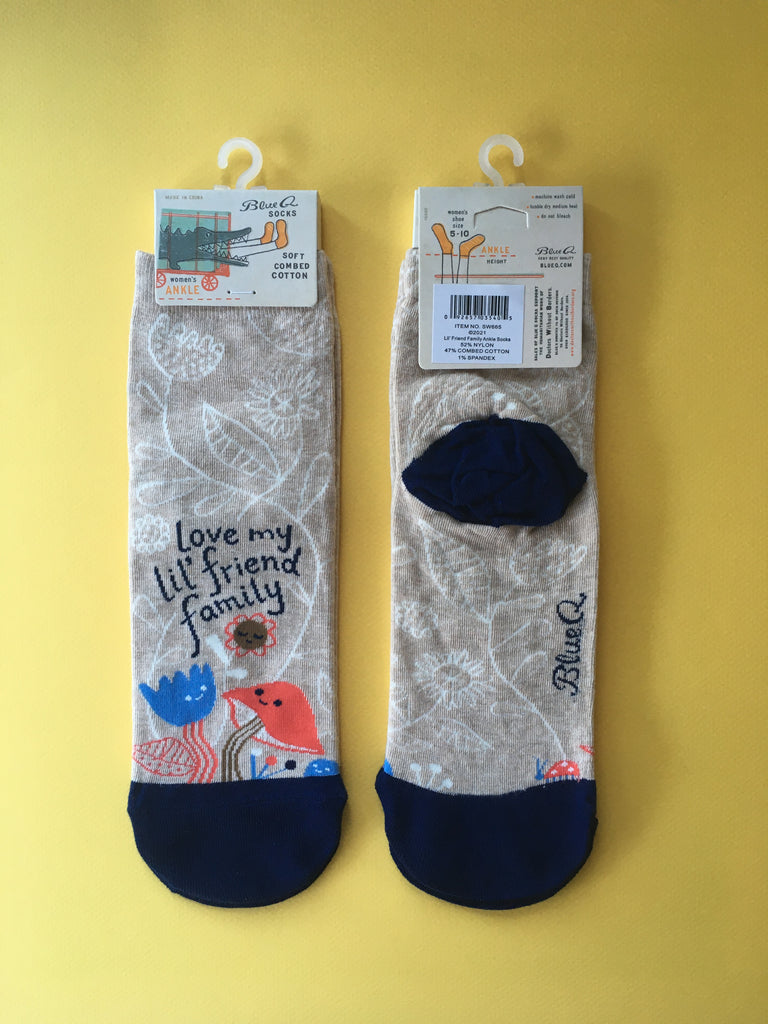 Overhead view of the front and back of the sock design laid flat against a yellow background. Socks are packaged with a Blue Q tab at the top. The socks are oatmeal colored with a navy blue toe. There's an illustrated image on the front of a mushroom, two flowers, and a rock with smiling faces all gathered up above the toe of the sock. Hand drawn type above them reads "love my lil' friend family". White linear floral pattern all over the background. Colors: Oatmeal, navy blue, bright sky blue, brown, bright red-orange, white.