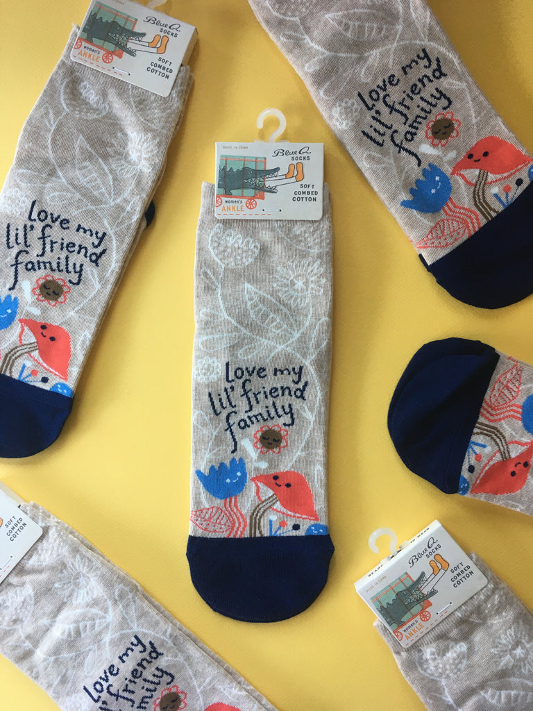 Overhead view of a bunch of the same sock design laid flat against a yellow background. Socks are packaged with a Blue Q tab at the top. The socks are oatmeal colored with a navy blue toe. There's an illustrated image on the front of a mushroom, two flowers, and a rock with smiling faces all gathered up above the toe of the sock. Hand drawn type above them reads "love my lil' friend family". White linear floral pattern all over the background. Colors: Oatmeal, navy blue, bright sky blue, brown, bright red-orange, white.