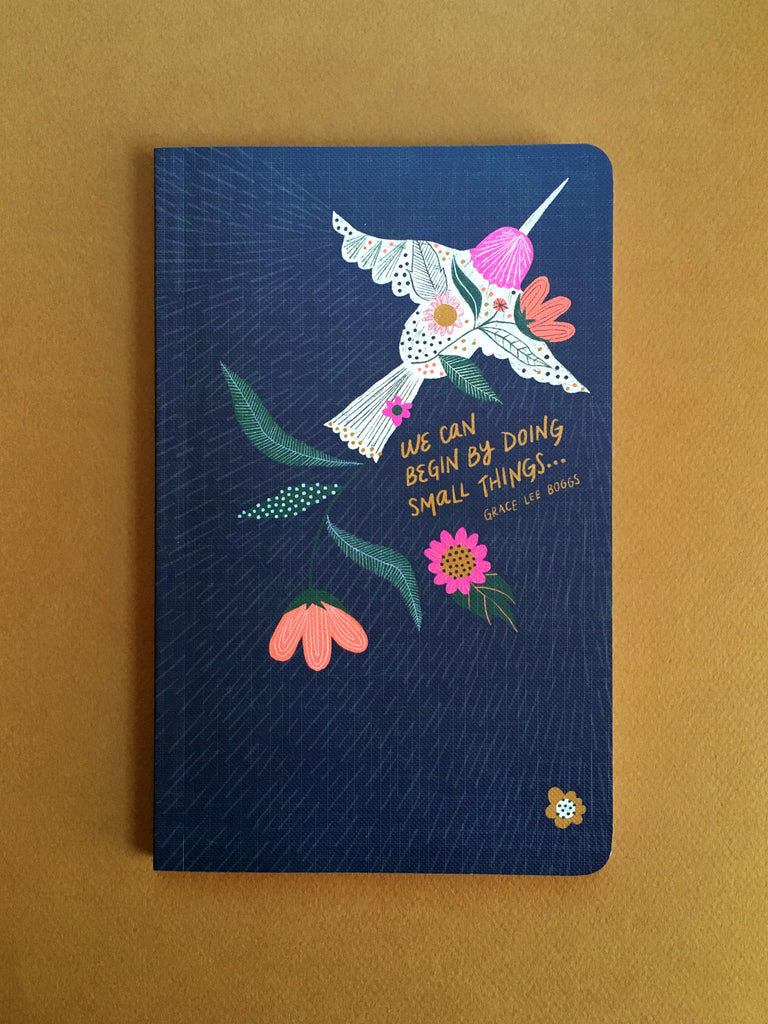 Straight on photo of the cover of the vertical journal. Dark blue-black background with a top view of a hummingbird flying towards the upper right corner and abstract flowers scattering behind it. 