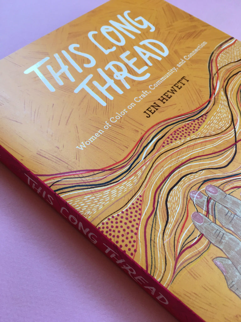 Angled closer up photo of a book with the title "This Long Thread" hand lettered in white at the top of the cover. Along the bottom of the cover are three illustrated hands and pattern-sleeved arms emerging up with the fingers intertwining with a horizontal band of threads, fibers, and stitches. Textural variations throughout along with dot and line patterns. Background color is a textural orange.