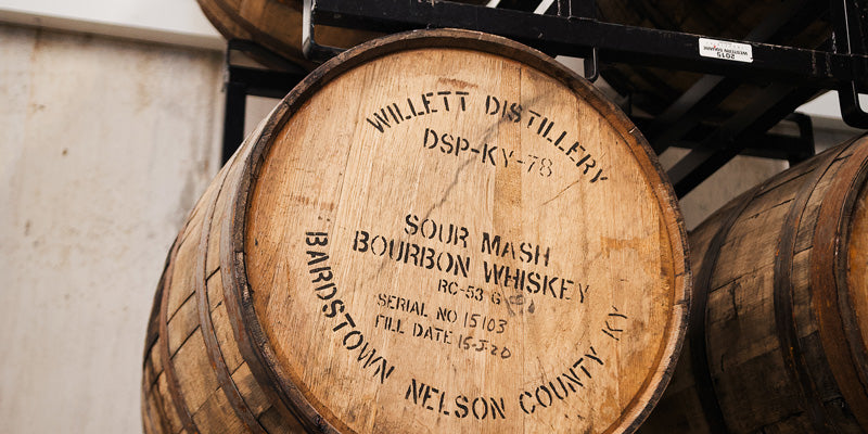 A Willett Bourbon barrel on a black steel rack with other barrels