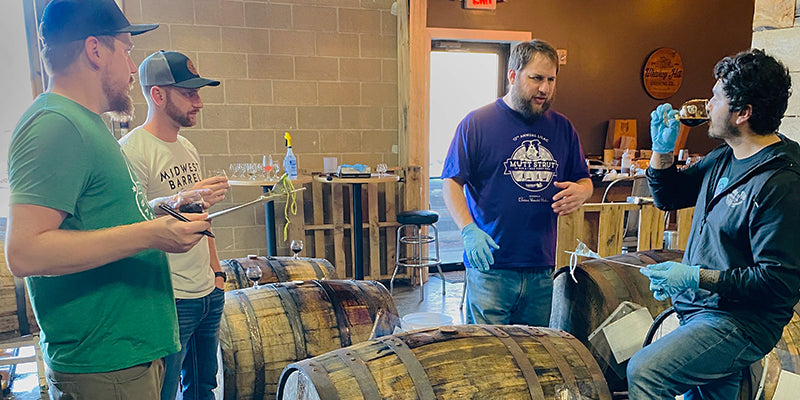 The Midwest Barrel Co. and Whiskey Hill Brewing teams standing near bourbon barrels, tasting samples and taking notes.