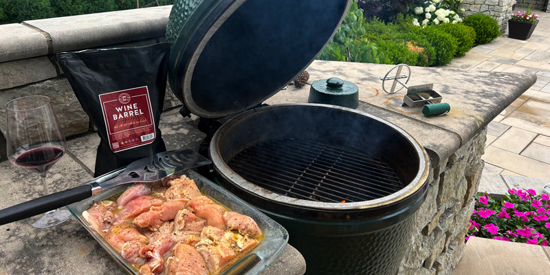 Raw chicken in a pan next to Midwest Barrel Co. Red Wine Barrel Smoking Wood Chips and a Big Green Egg charcoal grill in an outdoor patio