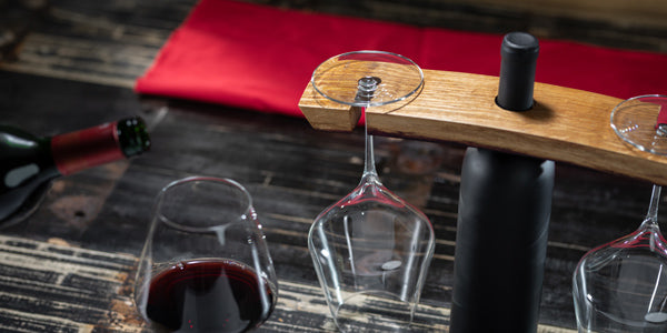 Glass of red wine being poured next to an unopened bottle and two glasses being held in a wine barrel stave holder