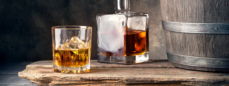 Tumbler glass of whiskey, next to decanter of whiskey and small barrel on top of a wooden tray