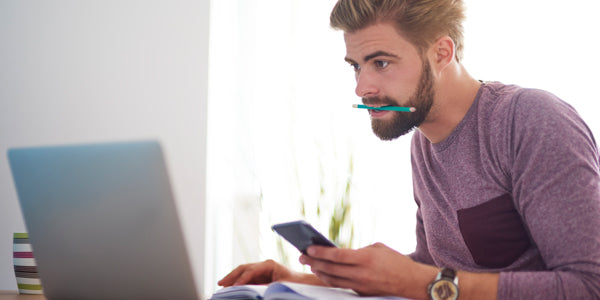 Man with pen in mouth, book open on lap, looking at laptop while holding calculator