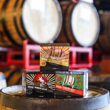Three boxed 4-packs of barrel-aged beer from Revolution Brewing placed on top of a bourbon barrel