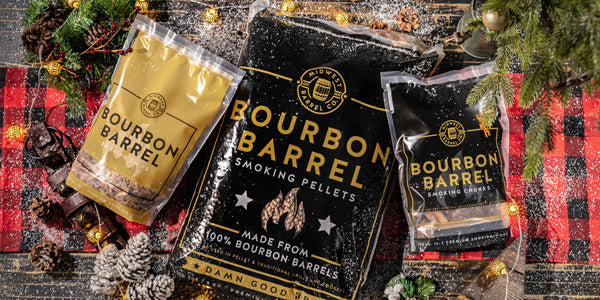 Bags of Bourbon Barrel Smoking Wood Pellets, Chips and Chunks on a red and black checkered blanket with pine needle branches, pine cones, snow and other Christmas themed decorations around the bags