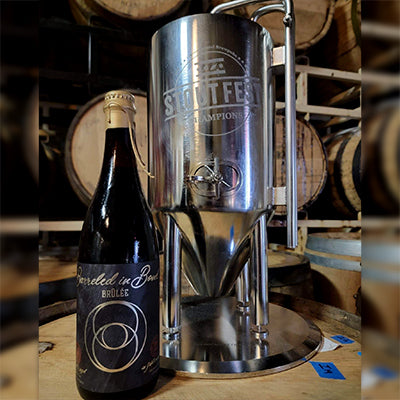 Bottle of Whiskey Hill Barreled in Bond barrel-aged beer next to a miniature beer tank trophy that says Stout Fest in front of filled bourbon barrels aging beer