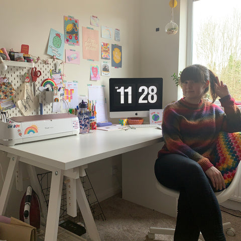 peach panda studio illustrator Rachel sat at her desk in her studio, wearing a colourful rainbow jumper surrounded by joyful stationery