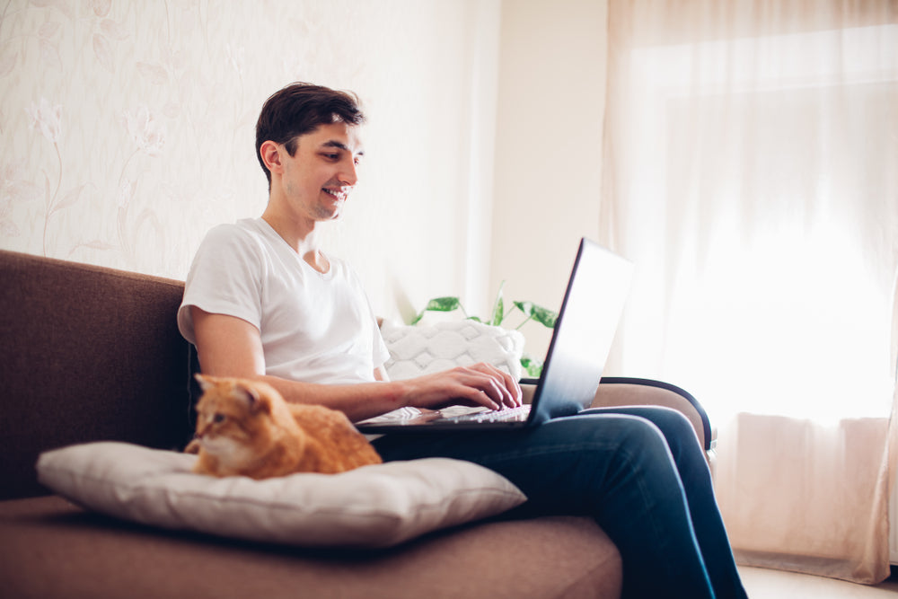 Man smiling at laptop during an online vet consultation, orange cat sitting next to him