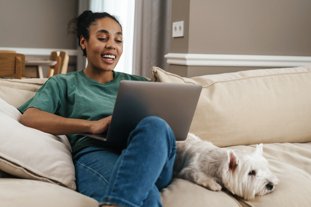 Young Black woman sitting on couch, smiling while on her laptop during virtual vet appointment; small white dog sitting next to her