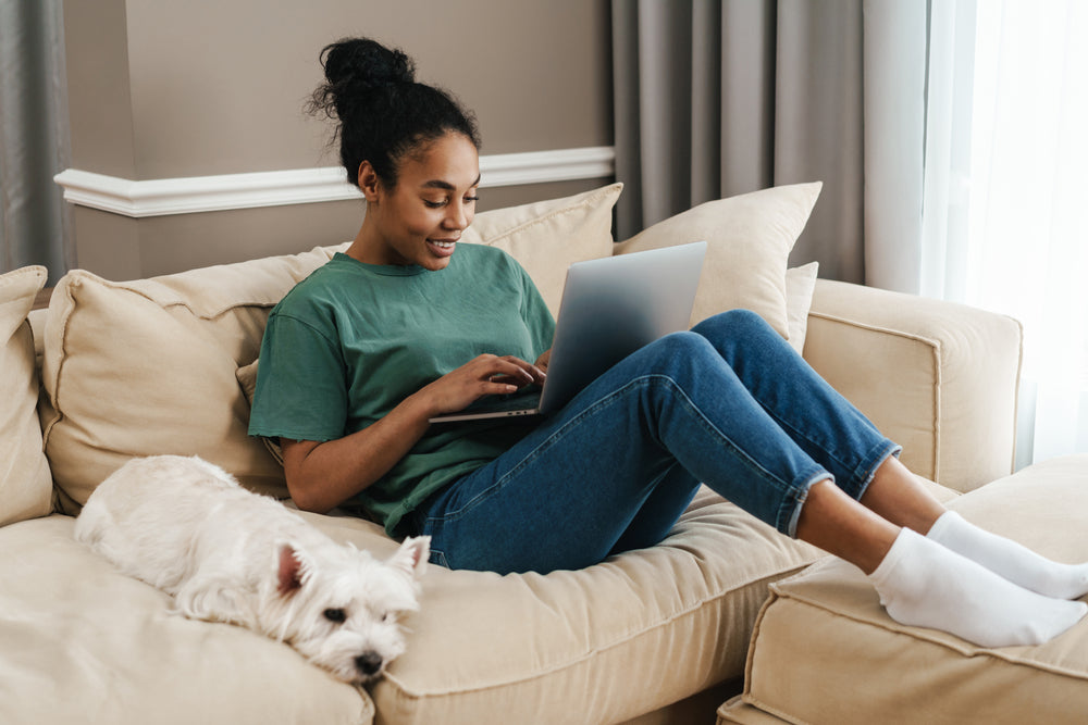 Young Black woman sitting on couch on her laptop waiting for an online vet appointment, white dog sitting next to her