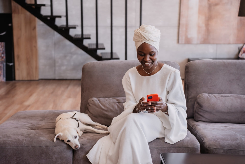 Black woman in turban looking at smartphone scheduling an online vet appointment for her dog sitting on couch next to her
