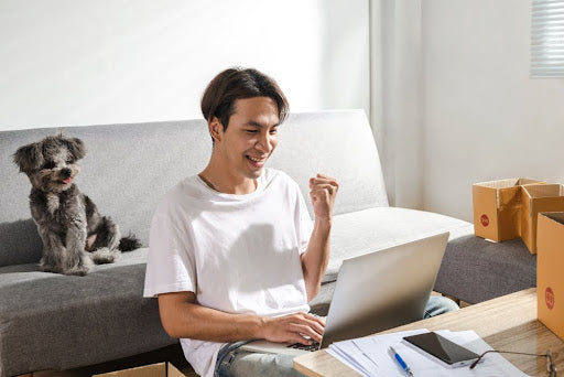 Young Asian man smiling during an online vet consultation; small dog sitting on couch behind them