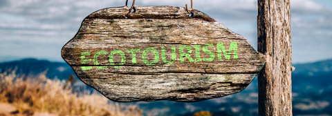 Wooden sign with 'Ecotourism' in green letters, with a background of mountains and tall grass, indicating a location dedicated to ecotourism