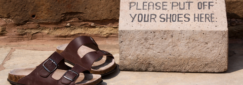 A pair of brown sandals next to a stone block with the words "Please out off your shoes here" written on it