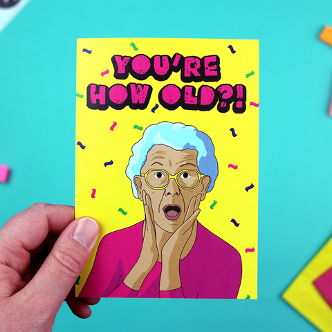 A hand holds a colourful card with an illustration os an older lady looking shocked under the words 'you're HOW old?!'