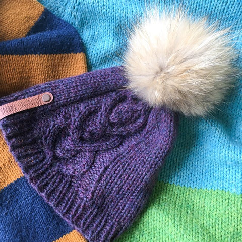 A purple cable knit hat with pom and navy & bronze striped scarf laid against a light blue and lime green sweater.