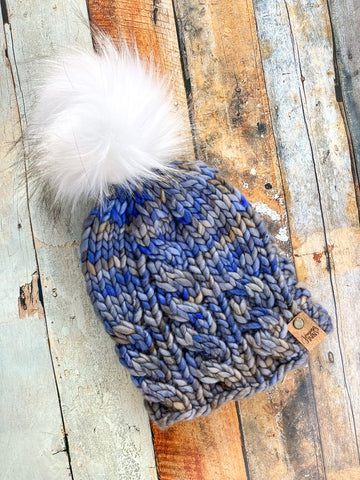 Cable knit beanie in light blue/gray variegated yarn with white pom against a wooded background.