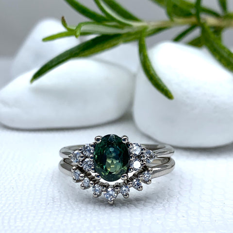 An oval green stone engagement ring sitting on top of a ring with a curve of diamonds that hugs the green gemstone. In the background are 3 white pebbles. In the top right corner a sprig of rosemary overlaps the pebbles.