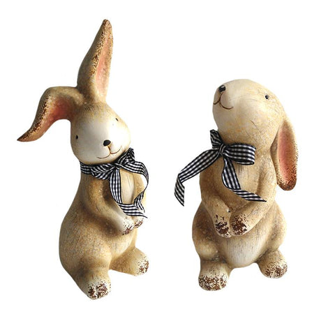 Two Bunnies standing one is looking forward over his shoulder and has his ears up the other is looking up with his ears laying down over his back.  Each is wearing a black plaid bow
