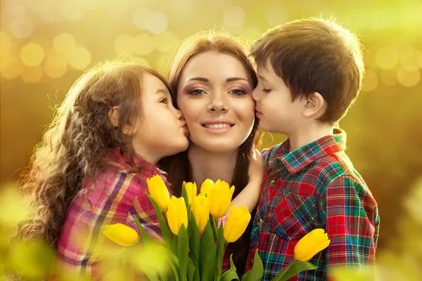 Woman with 2 children kissing her on both cheeks.  She is holding a bouquet of tulips.  In the distance there is a field of yellow.