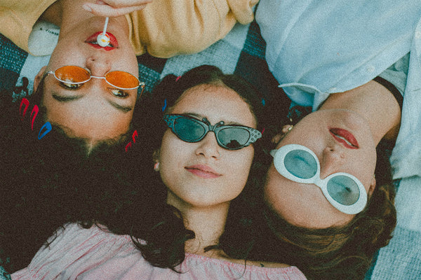 Overhead image looking down on the faces of three diverse women, laying on ground outside, looking at camera with sunglasses on.
