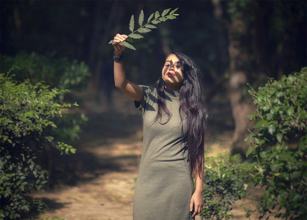 Image of woman standing outside, looking to sun with eyes closed, holding a branch of leaves to face to block sunlight