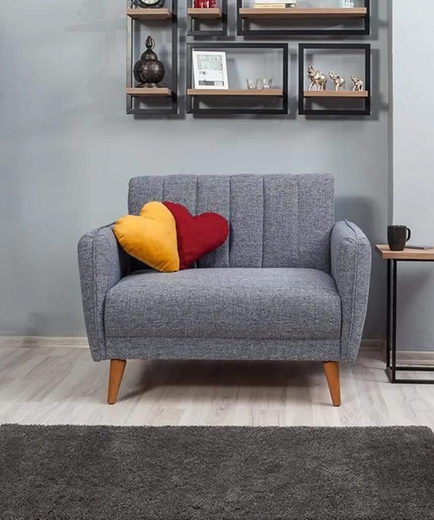 A grey wide single seater sofa, made with fabric. It has two little heart-shaped pillows on it in the colours of red and yellow.