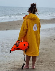 Orange swim buoy, women standing on beach in yellow changing robe holding swim buoy