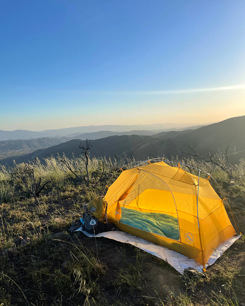 a yellow tent with a view of mountains behind it and the setting sun.