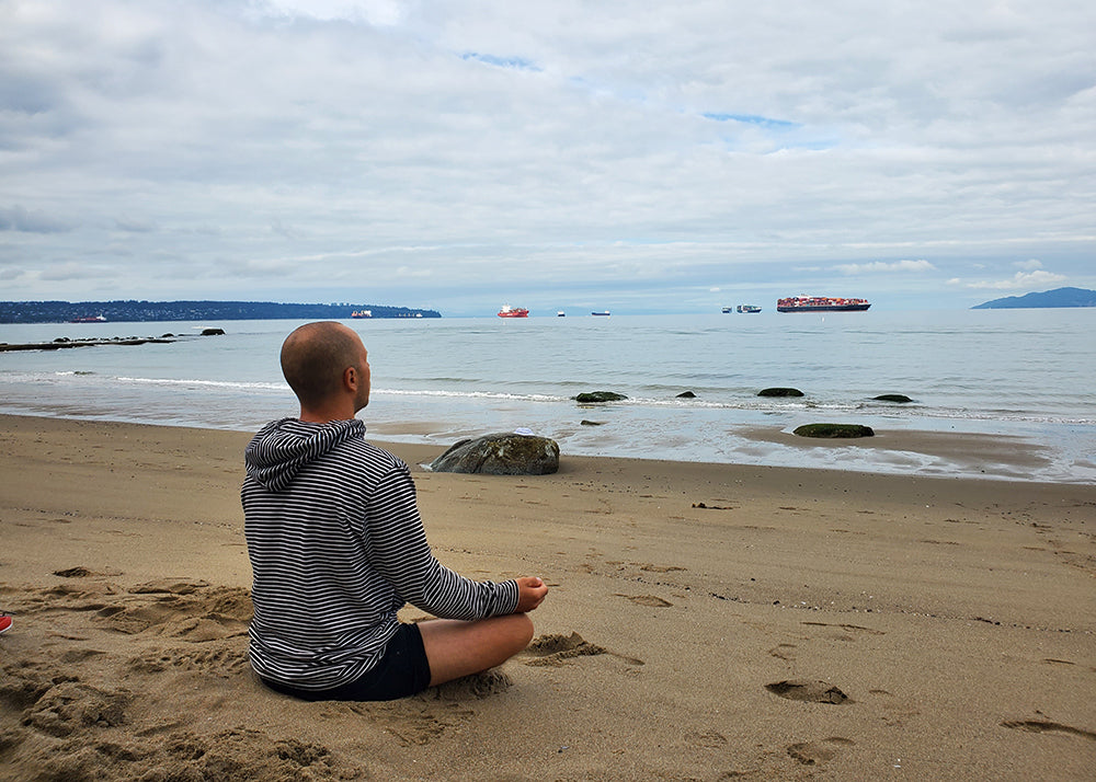 A man meditating on a beach looking out over a harbor with cargo ships in the distance