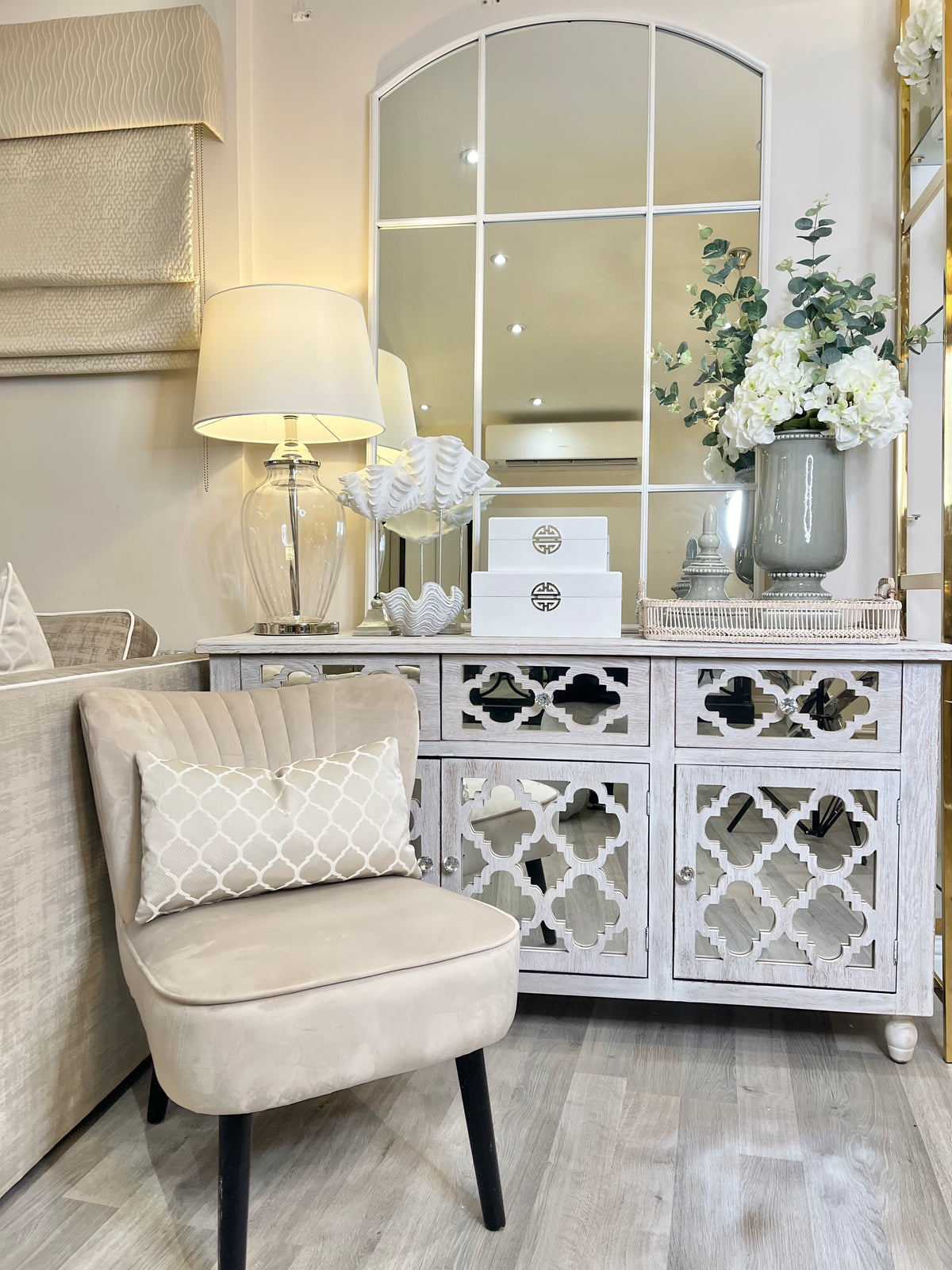 Wooden sideboard styled with ceramic accessories, silk flowers, glass table lamp and an arched window wall mirror 