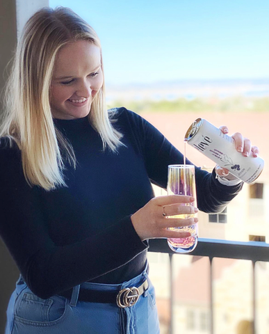 Blonde woman with medium to long hair wearing a black turtleneck with jeans. She pours a can of élevé sparkling water into a champagne glass, smiling.