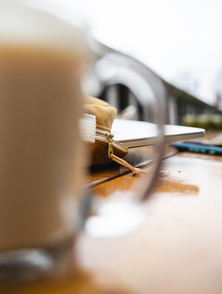 The corner of the backpack is visible through the handle of an out-of-focus glass coffee mug filled with a cappuccino