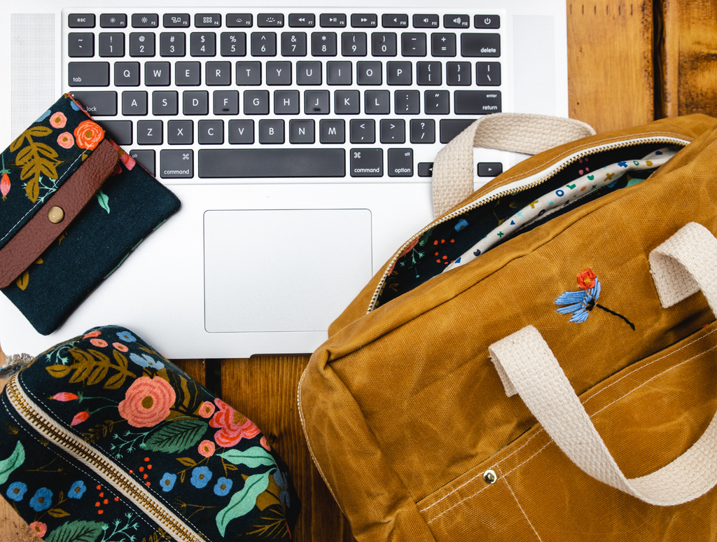 Flatlay of honey-colored waxed canvas backpack, laptop, Rifle Paper Co fabric pencil pouch, and mini wallet