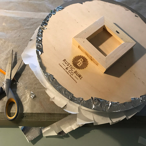 Pair of scissors on a table surface. Wooden circle with Rustic Burl & Co lettering visible.  Pieces of metal leaf adhered to the wood