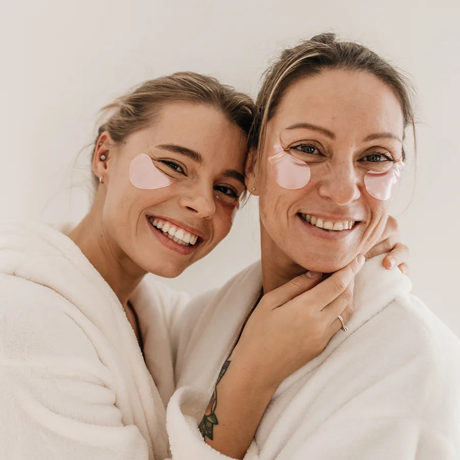 Deux femmes en peignoirs avec des patchs pour les yeux roses, se détendant lors d'un soin du visage.