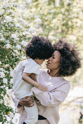 A black woman holding her son out playing in the park near a white rose brush or tree and very green all around.