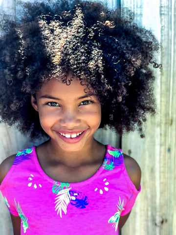 Multiracial child with super curly hair smiling in a very pink floral shit in-front of a door.