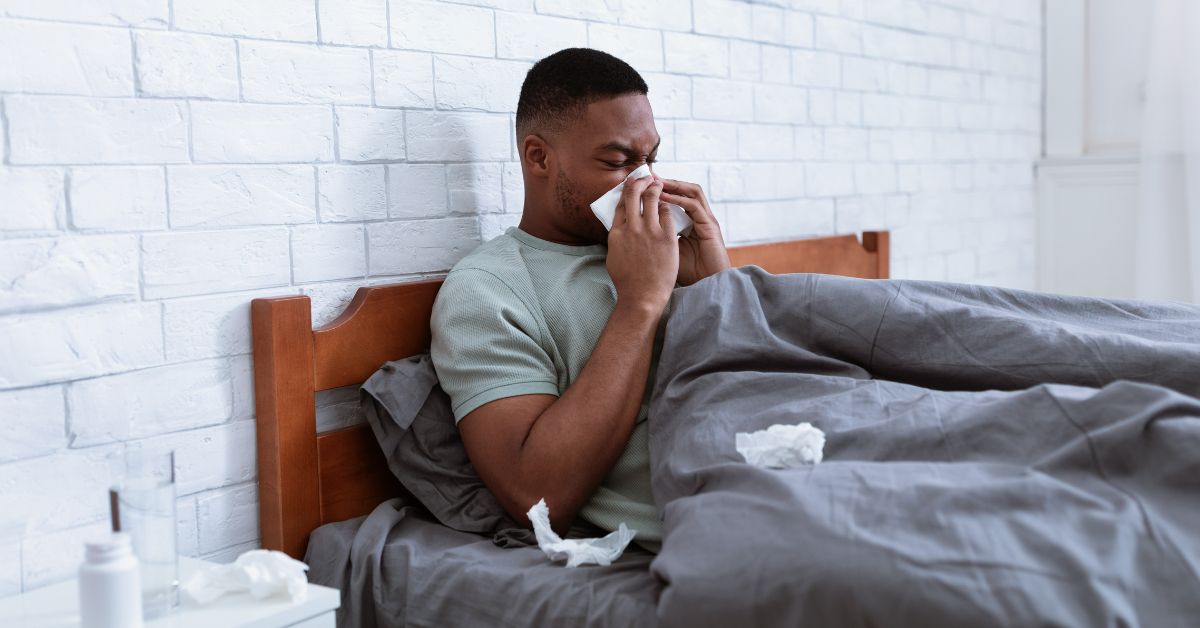 man blowing nose into tissue while lying in bed