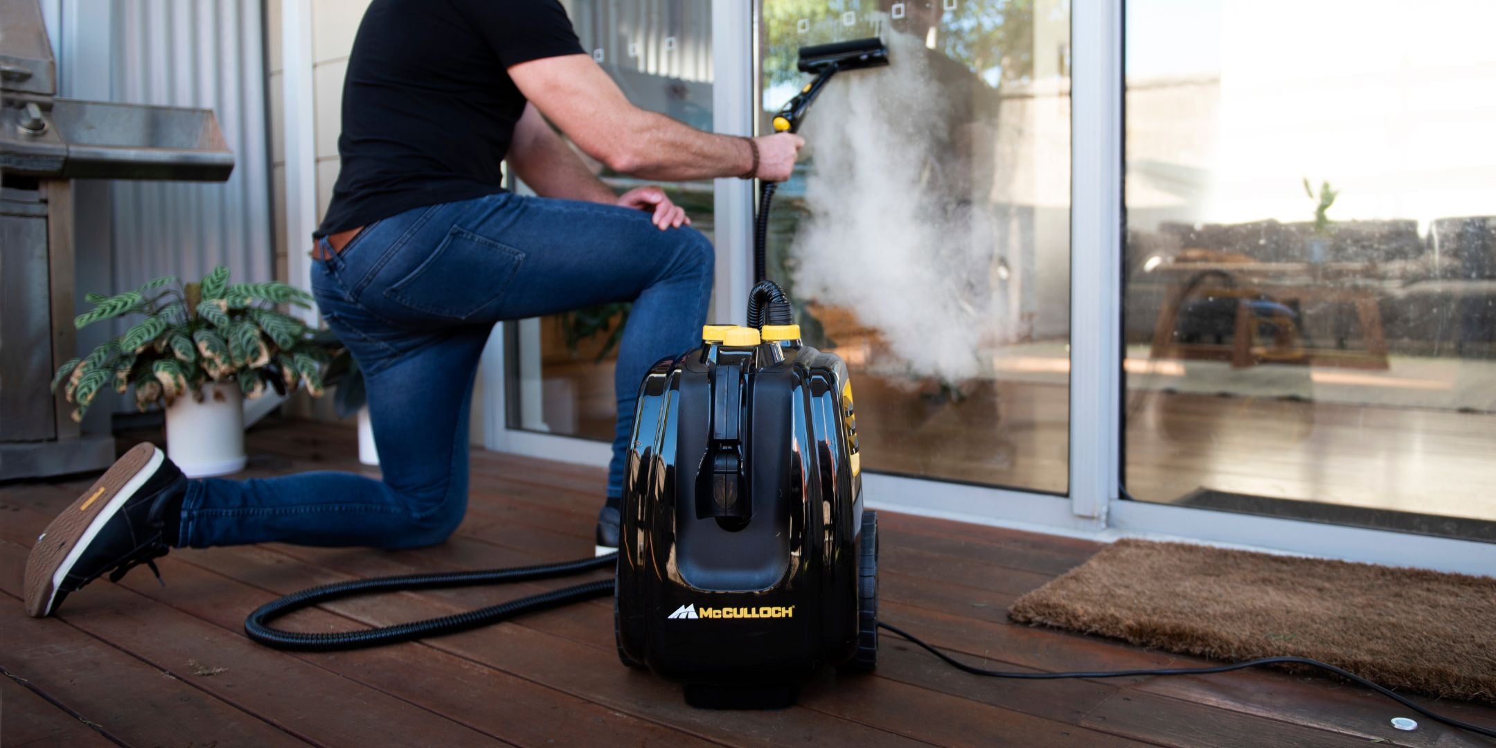 Man kneeling down to use McCulloch Outdoor & BBQ Steam Cleaner to steam clean window glass outside on the deck.