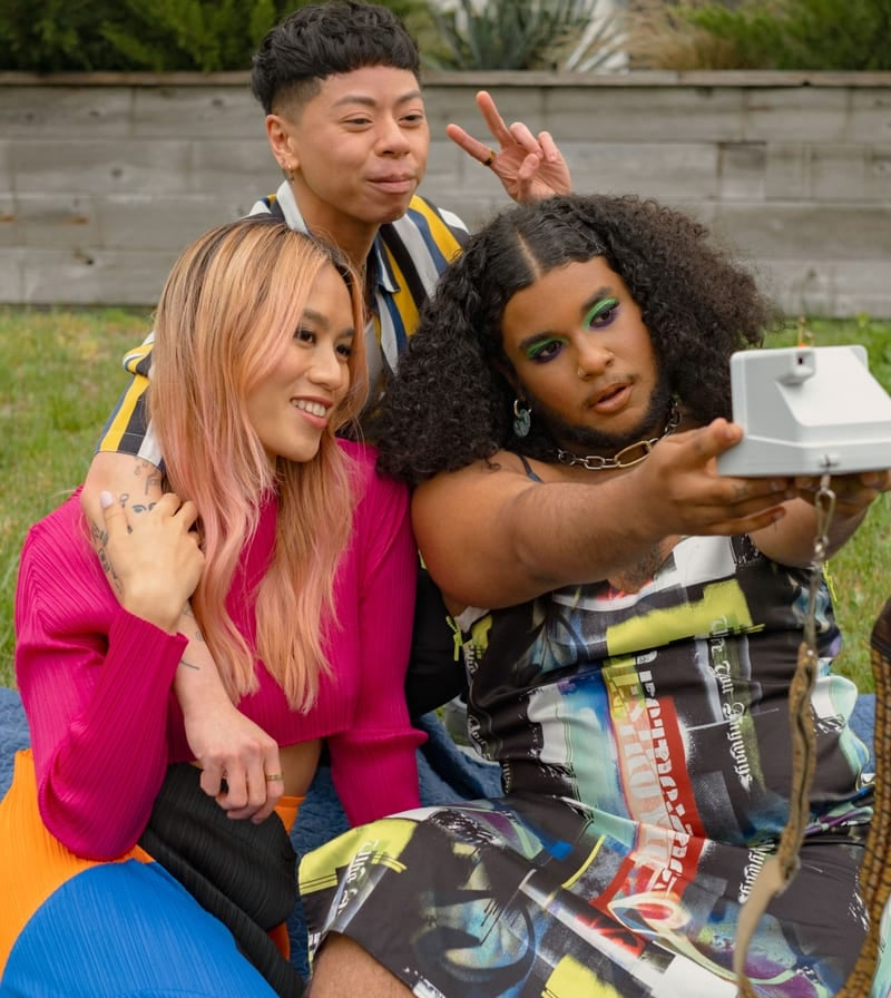 Three friends dressed in unique, patterned, and colorful clothing take a selfie while outside at a picnic together using a polaroid camera