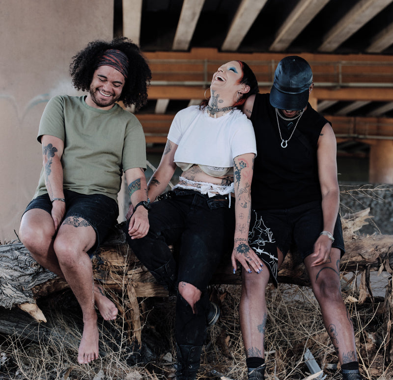 Three individuals all dressed in darker toned clothes laugh and hold hands while sitting underneath an industrial bridge