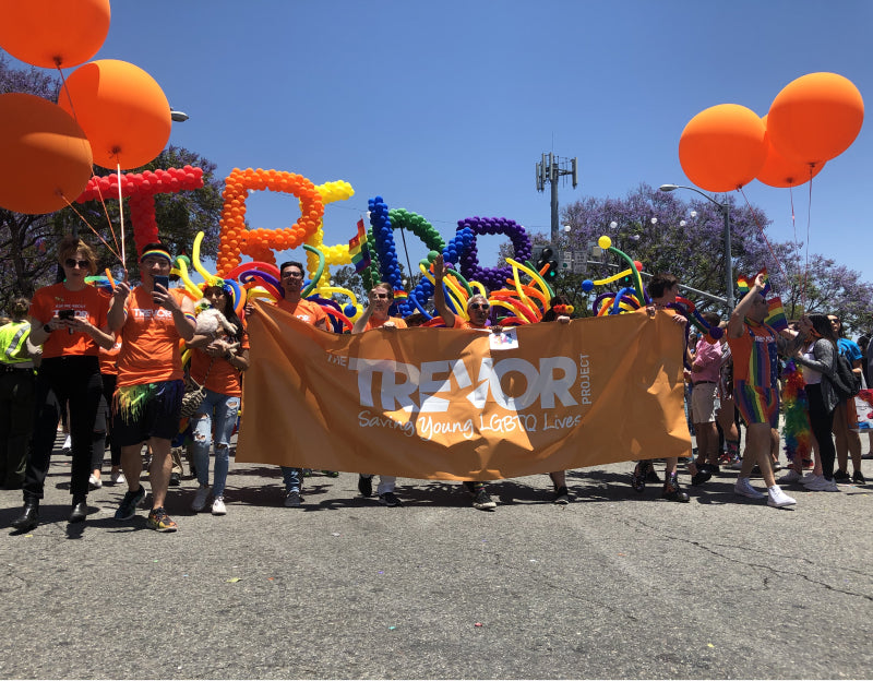 Members of the Trevor Project marching in an LGBTQ+ pride march holding a Trevor Project orange banner and holding rainbow ballons that spell out Trevor