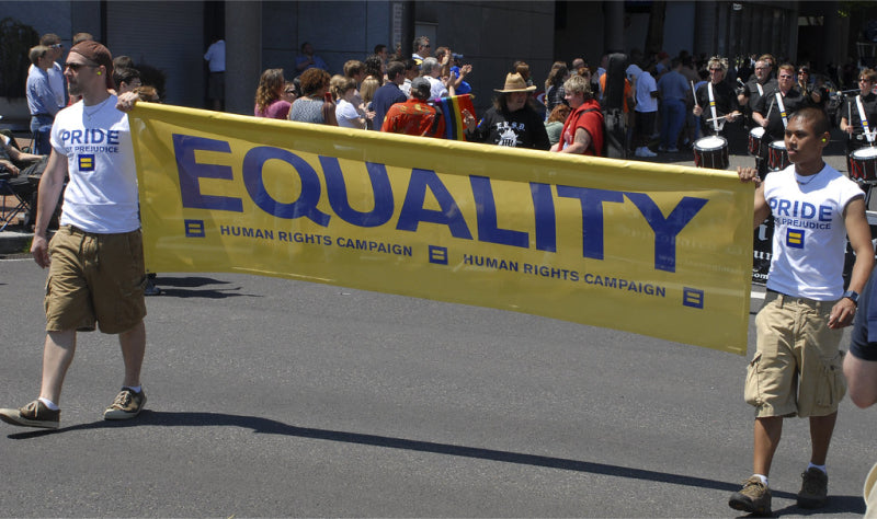 Two Human Rights Campaign members wearing pride t-shirts and holding up a banner that reads “EQUALITY” at a LGBTQ+ pride march