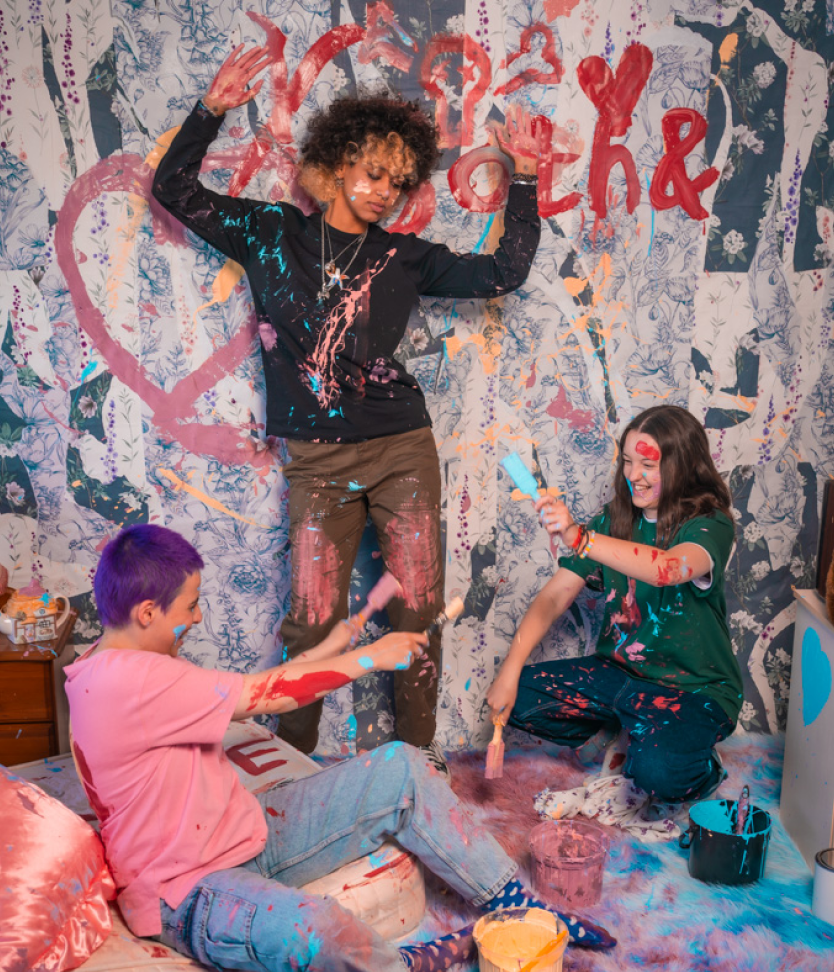 Photoshoot image of Bella Ramsey and two community members Bea and Nadia, in a colorful backdrop playing with paints