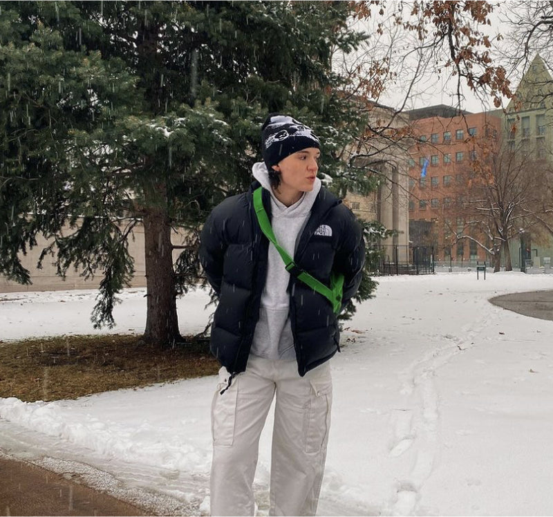 Masc person with short hair wearing a black and white beanie, black puffer jacket, and cream cargo pants with a green cross body purse